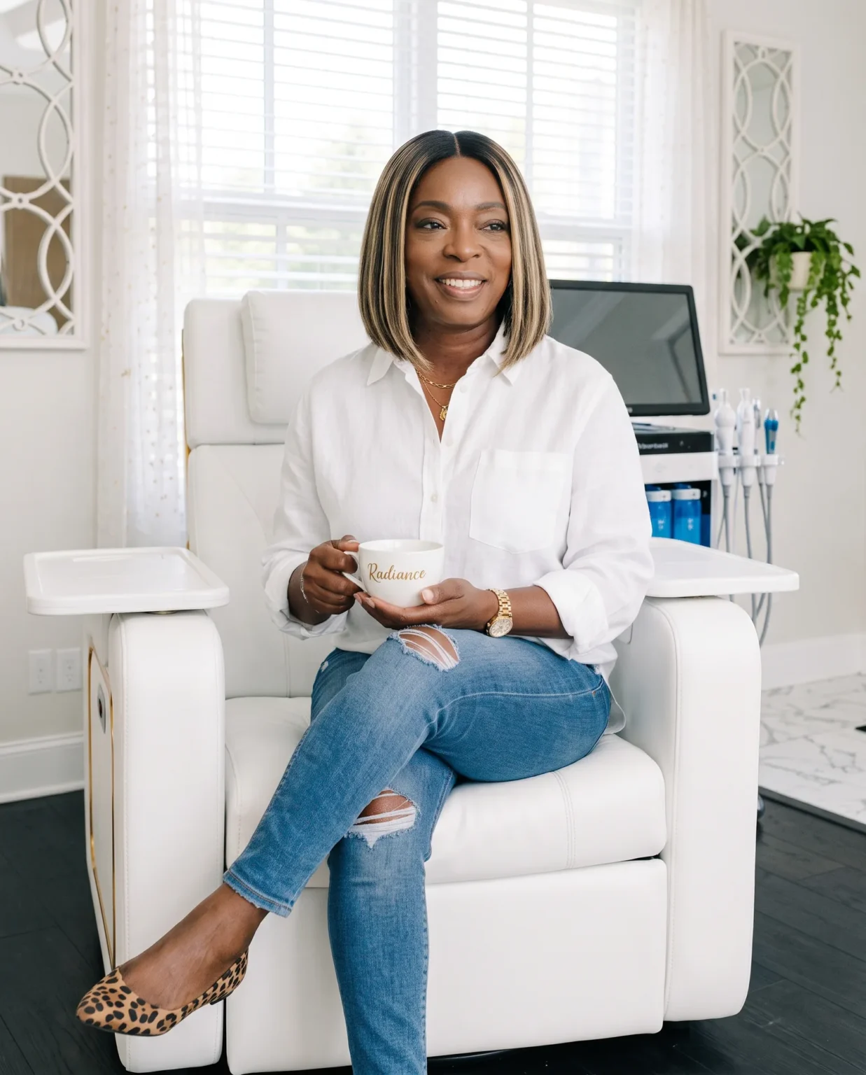 Portrait of Raytillia, owner of Radiance by Raytillia, smiling and seated in the studio’s white treatment chair, holding a Radiance coffee cup.